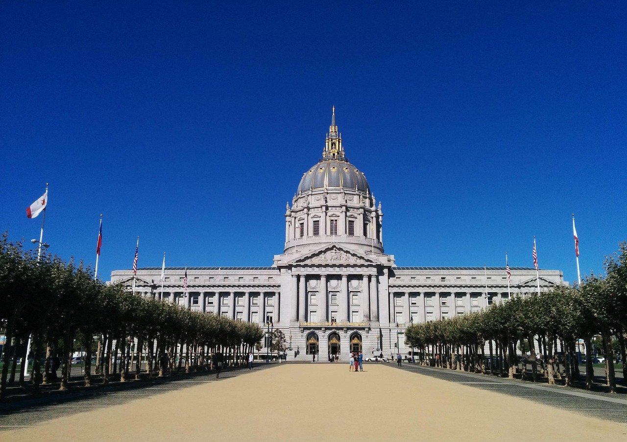 San Francisco City Hall med storslagen kupol och flaggor under klarblå himmel, omgiven av träd och öppen plats