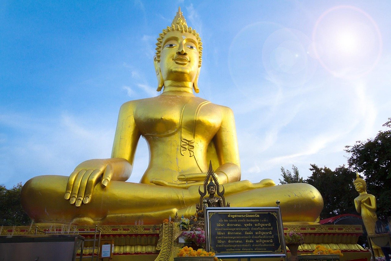Stor gyllene Buddha-staty sittande utomhus under blå himmel med mindre statyer och thailändsk skriftskylt framför