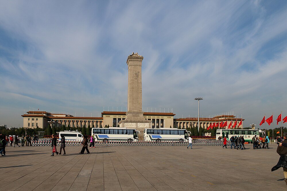 Himmelska Fridens Torg med Monument och Besökare Himmelska Fridens Torg i Peking med Monument till Folkets Hjältar, bussar, röda flaggor och turister under en blå himmel