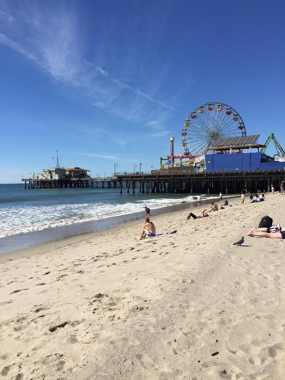 Solig strand med människor som kopplar av på sanden nära havet och den ikoniska Santa Monica Pier med pariserhjul under klarblå himmel