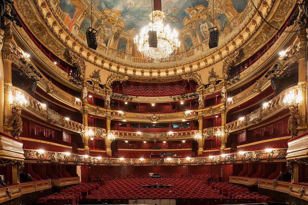 Interiörvy av en utsmyckad historisk teater med röda sammetsstolar, gyllene dekorationer och en stor ljuskrona