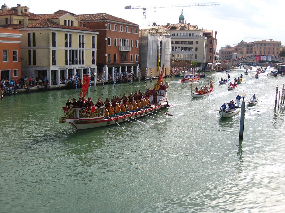 Venedigs kanalregatta med traditionella roddbåtar Traditionella roddbåtar med roddare i färgglada uniformer på en kanal i Venedig under ett regattaevenemang med historiska byggnader