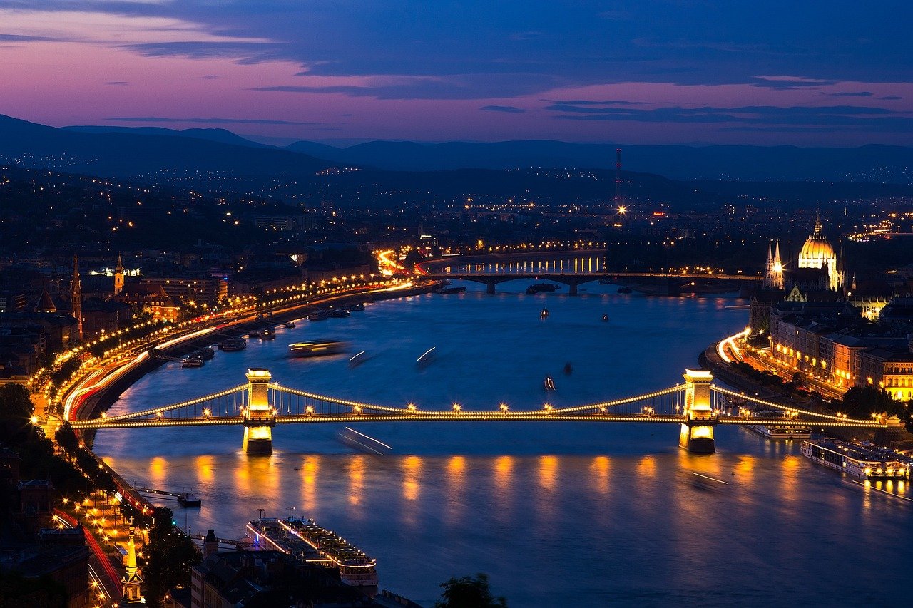 Nattvy av upplyst Kedjebron över Donau i Budapest med stadsljus och kullar i bakgrunden