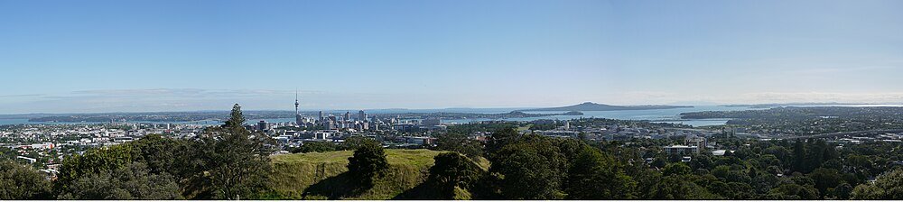 Aucklands stadssilhuett med Sky Tower och grönska Panoramavy över Aucklands stadssilhuett med Sky Tower, frodig grönska och omgivande vatten under klarblå himmel