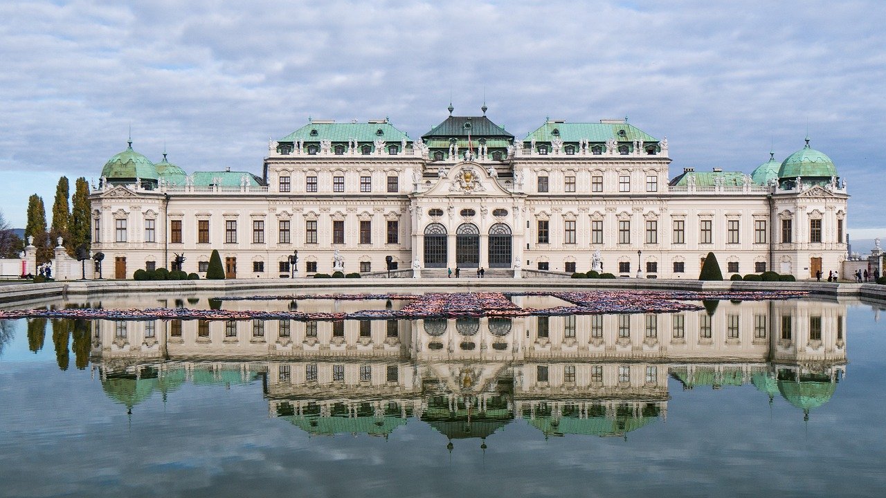 Barockpalats med gröna kupoler reflekterade i en stor damm under en molnig himmel, som visar historisk arkitektur.