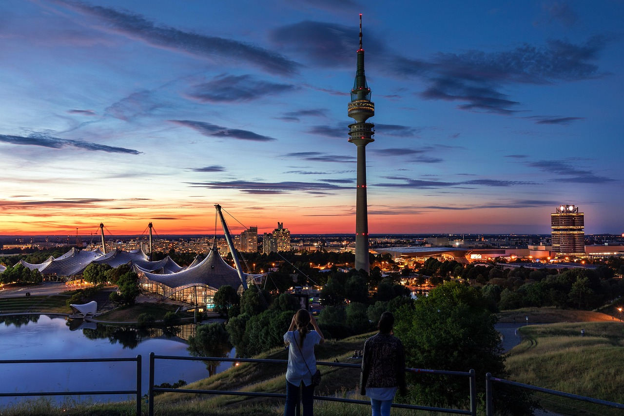 Münchens stadsbild med Olympiatornet vid solnedgången Solnedgångsvy över Münchens stadsbild med Olympiatornet och upplysta byggnader som reflekteras i vattnet