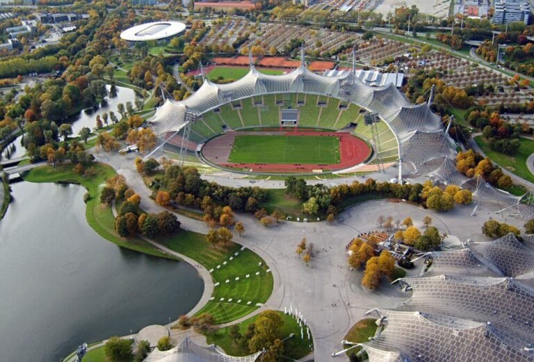 Flygfoto av Münchens Olympiastadion med sitt karakteristiska tälttak, omgivet av Olympiaparks sjöar och träd